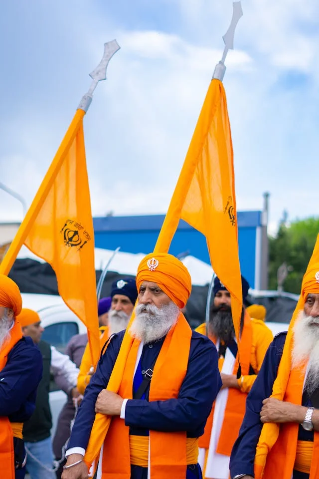 A colorful Nagar Kirtan procession for a Gurpurab celebration, with Sikhs carrying bright orange Nishan Sahib flags.