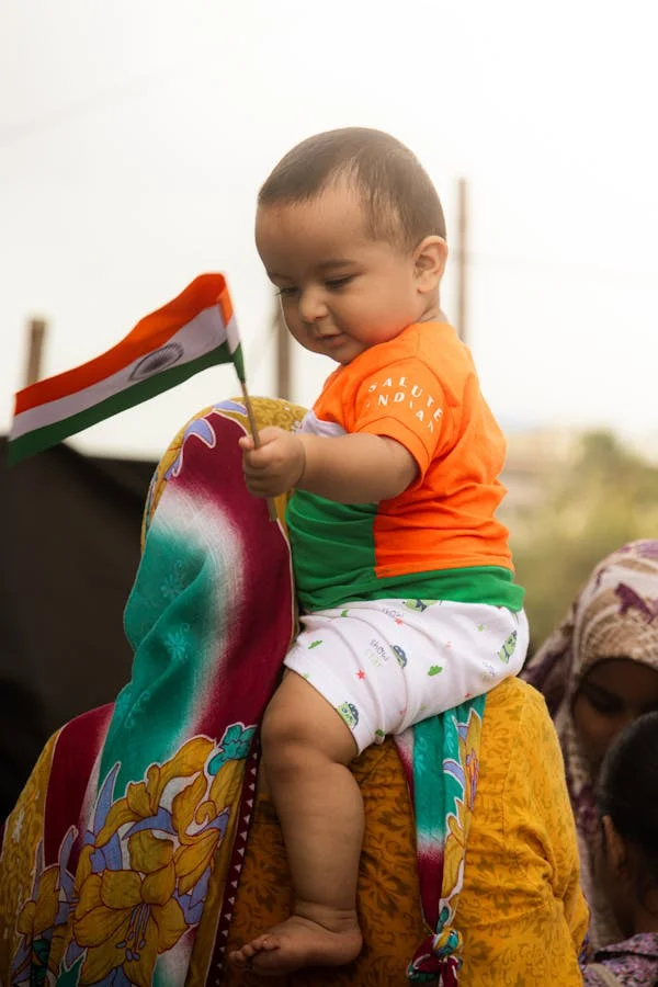 Baby in tricolor clothes holding Indian flag while sitting on shoulders during Independence Day celebrations.