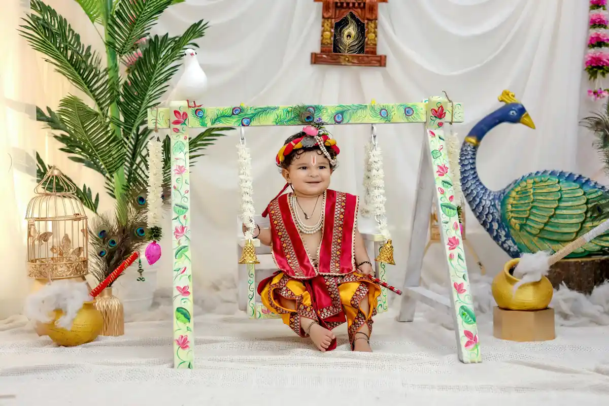 A young child dressed in costume as Bal Krishna (infant Lord Krishna) sits on a decorated swing as part of a family's Janmashtami celebration.